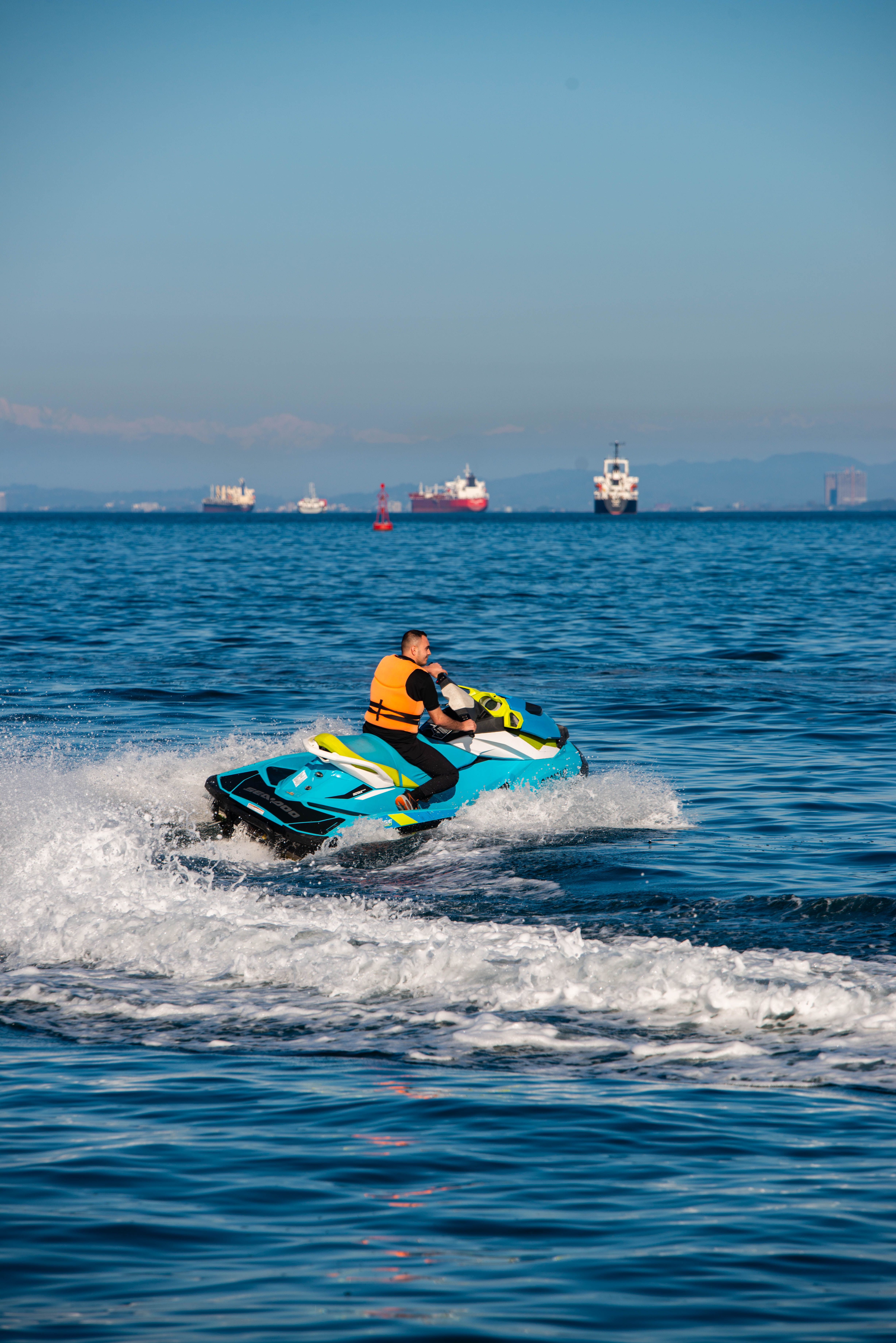 A rider on a personal watercraft wearing an orange life jacket, ships visible on the horizon